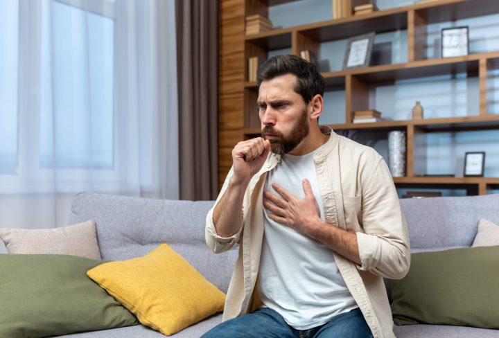 Sick mature man alone at home sitting on sofa coughing holding hands to chest in living room.