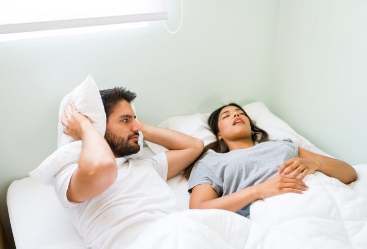Upset young man covering his ears with a pillow and feeling angry with his snoring girlfriend while lying in bed